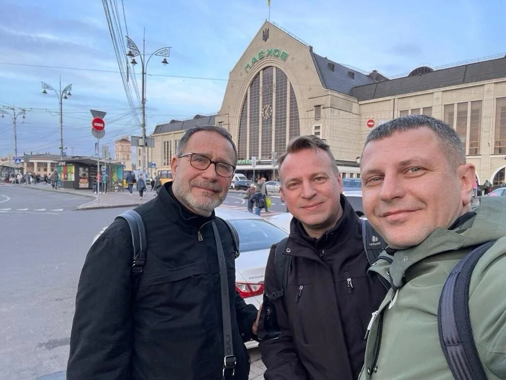 Trois hommes devant une gare animée.
