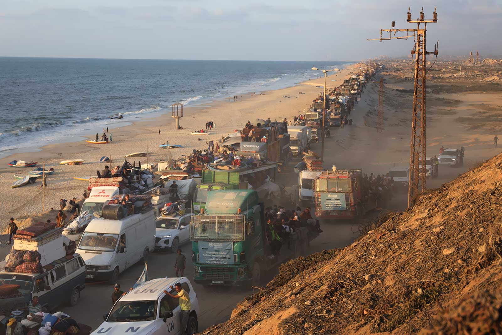 Long convoi de familles déplacées et de camions chargés sur la route côtière de Gaza, au crépuscule.
