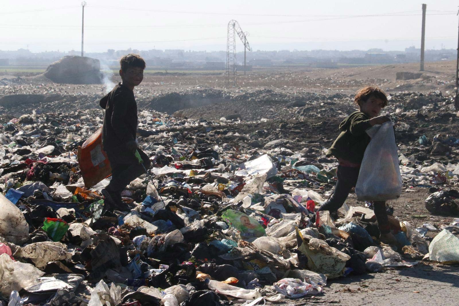 Deux enfants ramassent des objets dans une décharge à ciel ouvert, aux abords d'une ville, un sac de plastique à la main.