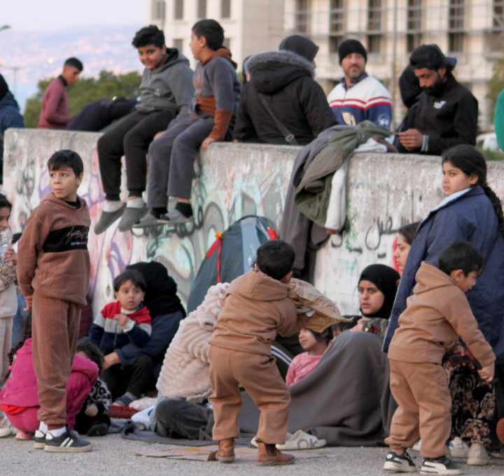 Des familles déplacées — enfants, femmes et hommes — installées le long d'un mur de béton, avec une tente et des affaires posées à même le sol.