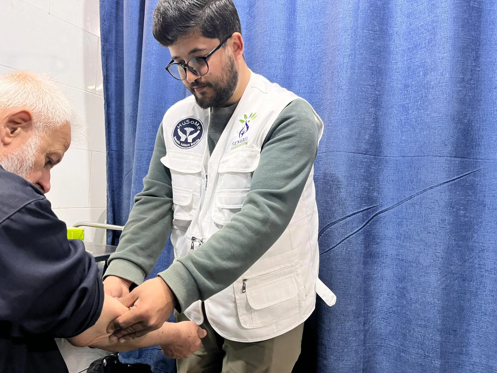 Un soignant en gilet HuSoMe et Senabil examine le bras d'un patient âgé, derrière le rideau d'un box de soins.