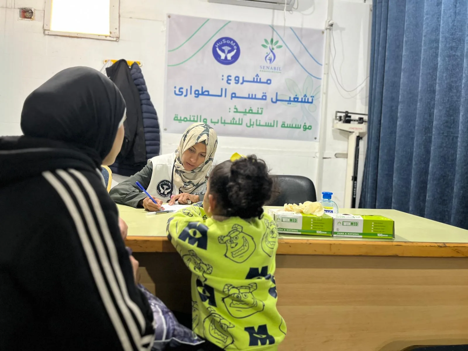 Consultation d'une enfant par une soignante, sous la banderole du projet de fonctionnement du service des urgences mené avec la Fondation Senabil.