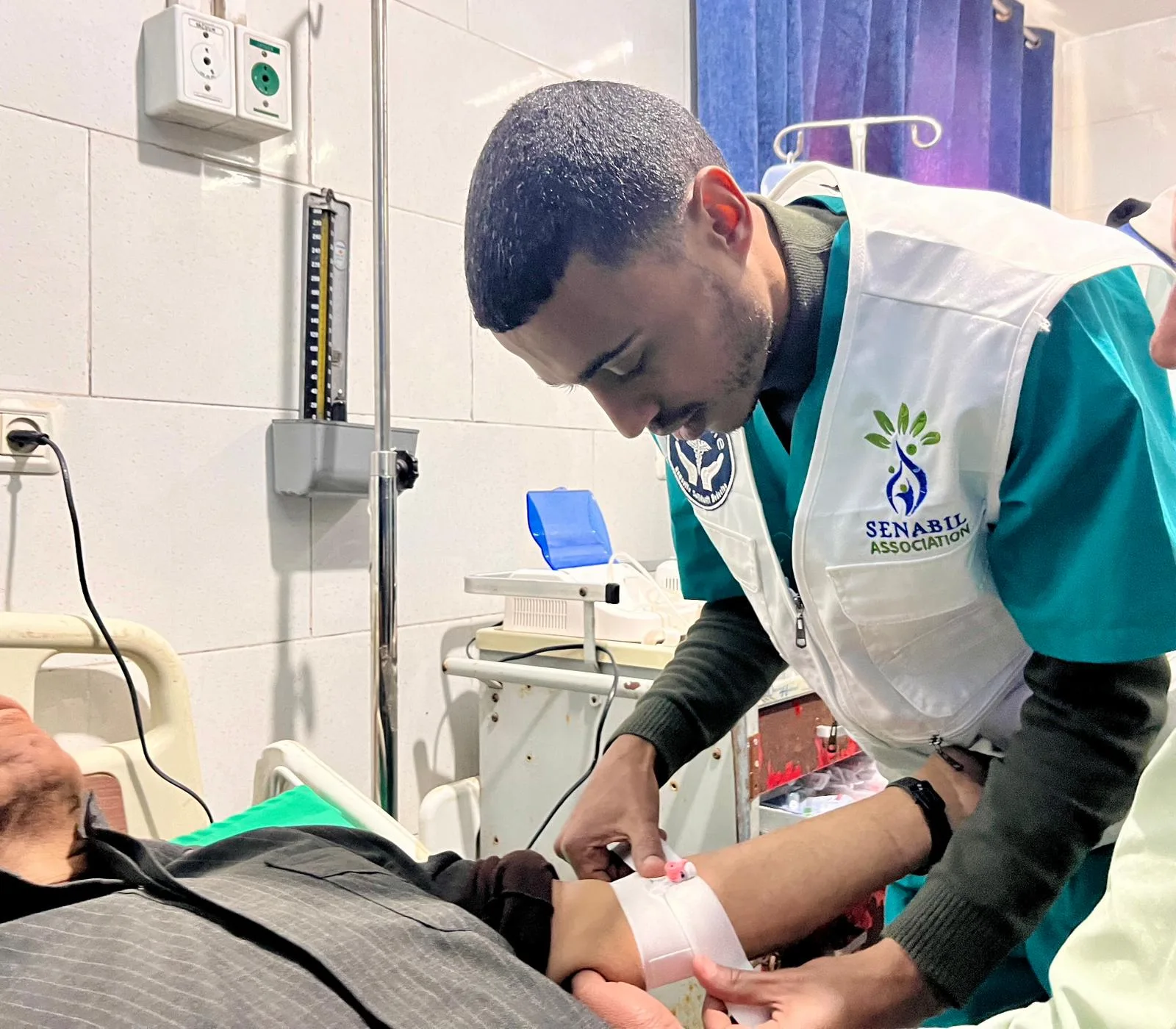 Un infirmier pose une perfusion au bras d'un patient allongé, dans une salle de soins.