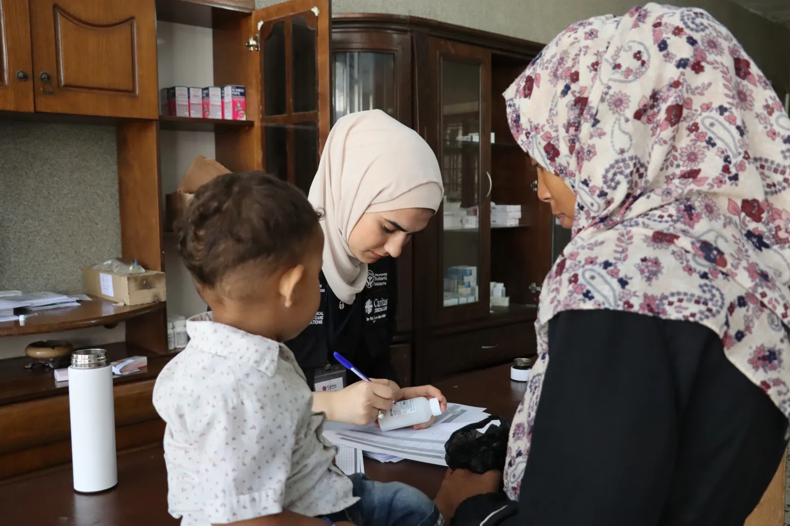 Une soignante rédige une ordonnance devant une mère et son jeune enfant, dans une salle dont les armoires vitrées contiennent les médicaments.