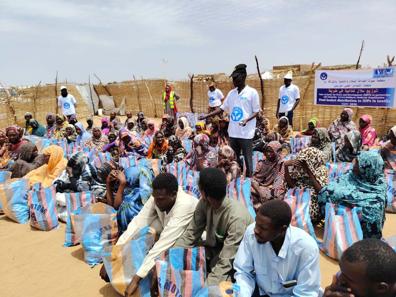 Distribution de colis alimentaires : des dizaines de familles assises dans le sable, chacune devant son sac, sous une banderole de la Voice of Justice for Peace and Development et de HuSoMe.
