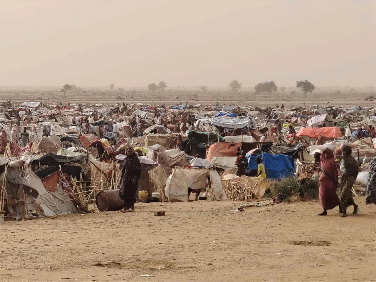 Un camp de déplacés au Darfour : des milliers d'abris de bâches et de branchages sur une plaine de sable, sous un ciel chargé de poussière.