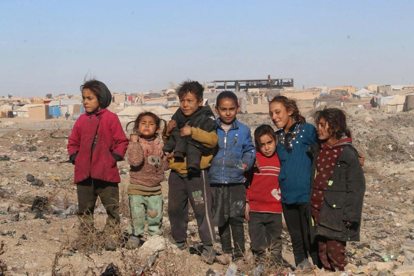 Un groupe d'enfants réunis à l'entrée d'un camp de déplacés, dans le nord-est syrien ; derrière eux, les abris de fortune s'étendent jusqu'à un bâtiment éventré.